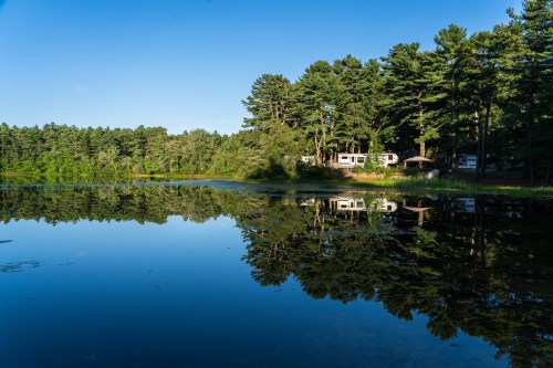A serene lake reflecting trees and cabins under a clear blue sky.