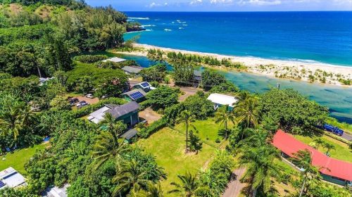 Aerial view of a coastal area with lush greenery, houses, and a sandy beach along a blue ocean.