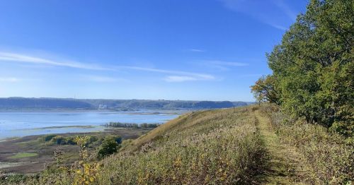 A scenic view of a river valley with a clear blue sky, surrounded by green hills and a walking path.
