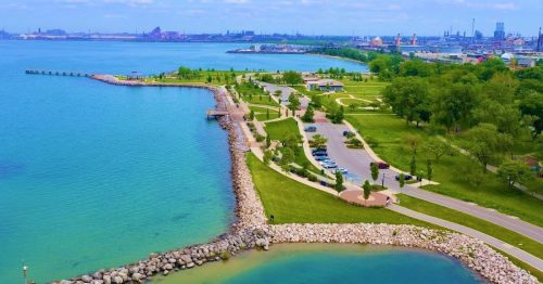 Aerial view of a scenic waterfront park with green spaces, pathways, and a calm blue lake. Industrial area in the background.