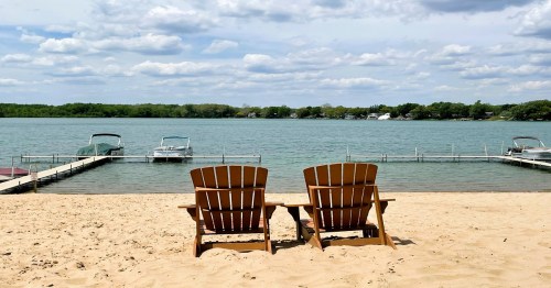 Two wooden chairs on a sandy beach facing a calm lake with boats docked and a cloudy sky above.