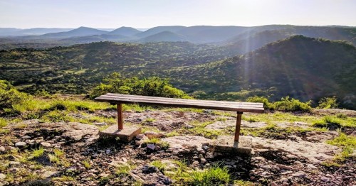 A wooden bench on a rocky outcrop overlooking a lush green valley and distant mountains under a clear sky.