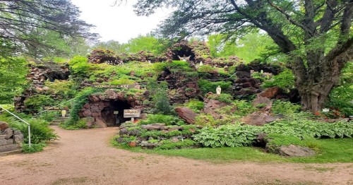 A lush, green garden featuring a rocky structure with an entrance, surrounded by trees and vibrant foliage.