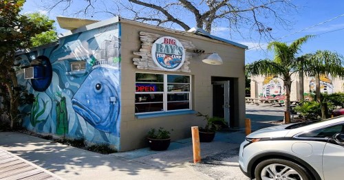 A small restaurant with a fish mural, "Big Ray's Fish Camp," featuring outdoor seating and palm trees.