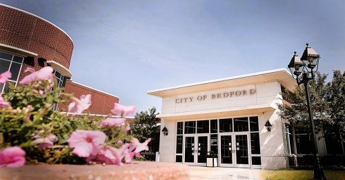 City of Bedford building with a modern facade, surrounded by flowers and lampposts under a clear blue sky.