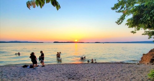 Sunset over a calm lake with people swimming and enjoying the beach, surrounded by trees and distant hills.