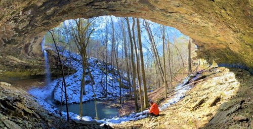A person in an orange jacket sits inside a cave, overlooking a snowy landscape with a waterfall and trees.