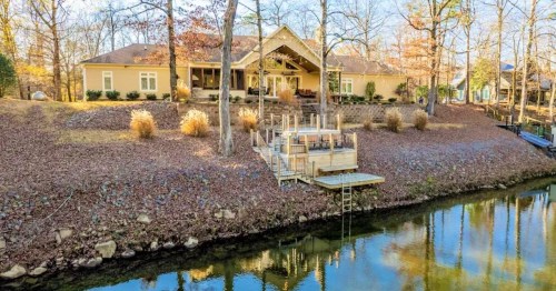 A serene lakeside home surrounded by trees, with a wooden deck extending over the water and autumn foliage.