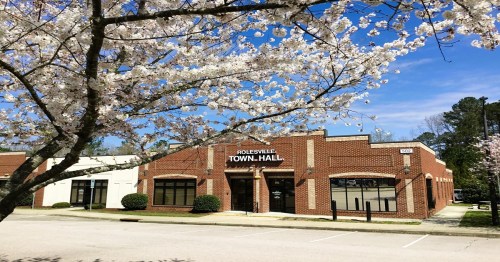 A brick building labeled "Rolesville Town Hall" surrounded by blooming cherry blossom trees and a clear blue sky.