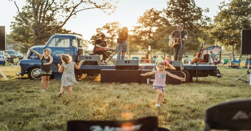 Children dance in a grassy field while a band performs on stage, with a vintage blue truck in the background.