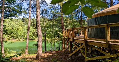 A wooden deck leads to a yurt by a serene green lake, surrounded by tall trees and lush foliage.