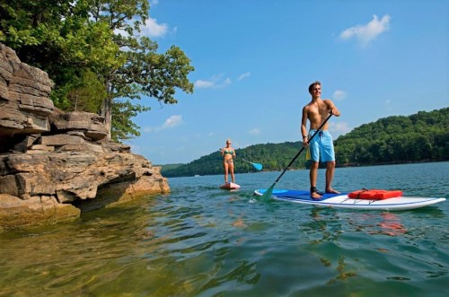 Two people paddleboarding on a calm lake, surrounded by trees and rocky cliffs under a clear blue sky.
