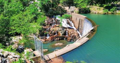 A serene waterfall cascades over a stone barrier into a calm green lake, surrounded by lush greenery.