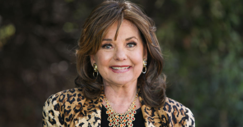 A smiling woman with brown hair, wearing a leopard print jacket and statement necklace, stands outdoors.