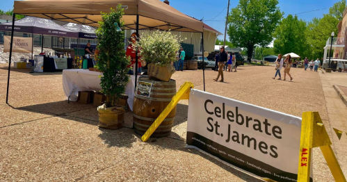 A festive outdoor event with a tent, barrels, and a sign reading "Celebrate St. James," surrounded by people and greenery.