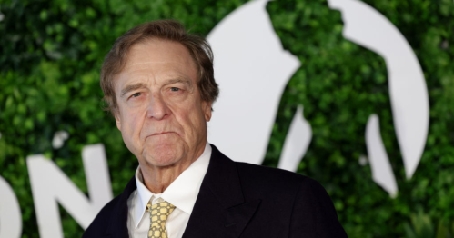 A man in a suit stands in front of a green backdrop with a silhouette, looking serious and confident.