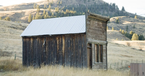 A weathered wooden shed stands alone in a grassy field, surrounded by rolling hills and sparse trees.