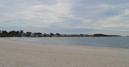 A serene beach scene with soft sand, calm water, and a distant shoreline under a cloudy sky.