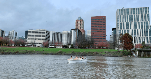A small boat with people floats on a river, with a city skyline and green park in the background.