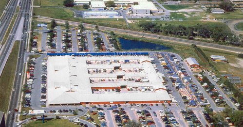 Aerial view of a large shopping center with parking lots, nearby roads, and green spaces.