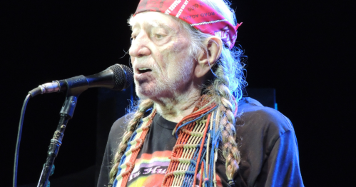 An elderly man with long hair and braids sings into a microphone, wearing a red bandana and a colorful shirt.