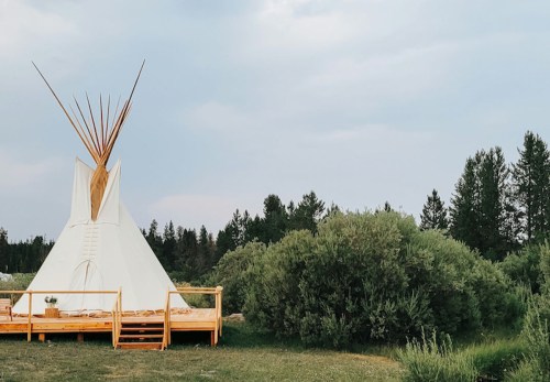 A white teepee on a wooden deck surrounded by greenery and trees under a cloudy sky.