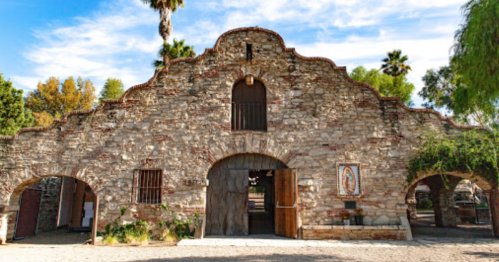 Historic stone building with arched entrance, surrounded by palm trees and greenery, featuring a religious image on the wall.