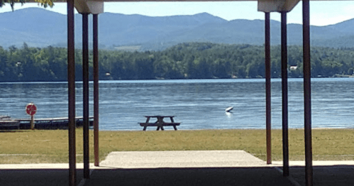 View of a calm lake with mountains in the background, framed by a structure, featuring a picnic table by the water.