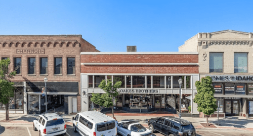 A street view of a historic downtown area featuring shops, trees, and parked cars under a clear blue sky.