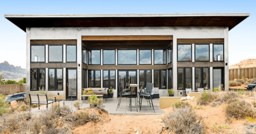 Modern house with large windows, surrounded by desert landscape and mountains in the background. Outdoor seating area visible.