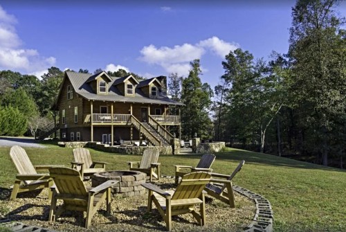 A large wooden house with a porch, surrounded by trees, and a fire pit with chairs in the foreground.