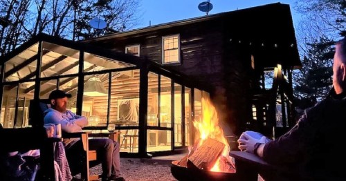 A cozy outdoor scene with a fire pit, two people relaxing, and a modern cabin illuminated at dusk.