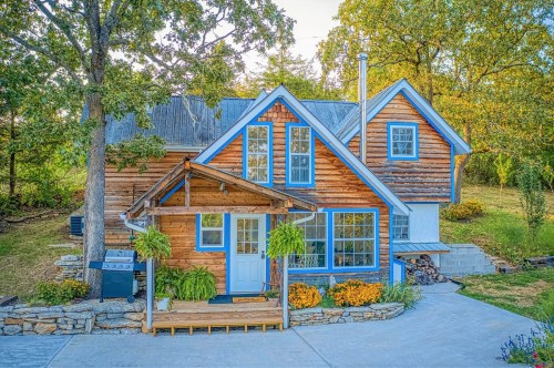 A charming wooden house with blue accents, surrounded by greenery and colorful flowers, featuring a welcoming porch.