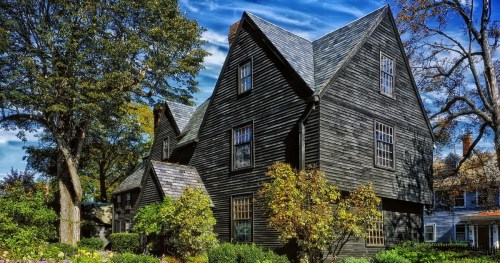 A large, dark wooden house with a steep roof, surrounded by trees and colorful foliage under a blue sky.