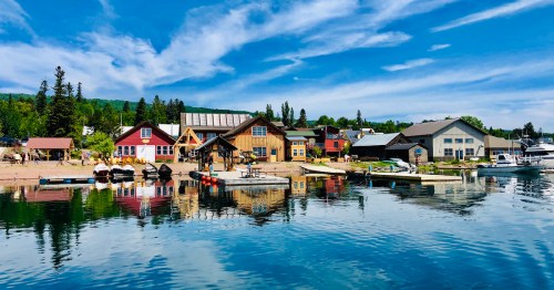 Colorful houses line a calm waterfront, reflecting in the water under a blue sky with scattered clouds.