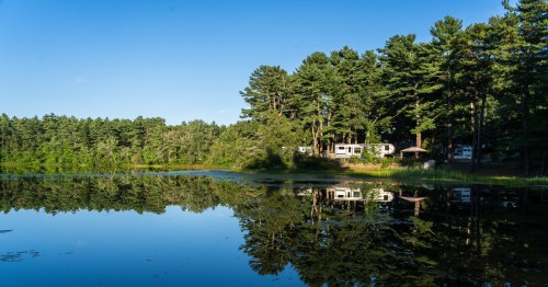 A serene lake reflects trees and a camper in a peaceful natural setting under a clear blue sky.