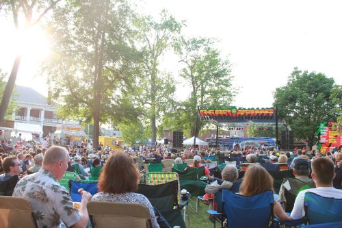 A lively outdoor concert scene with a large crowd seated on lawn chairs, enjoying a performance under sunny skies.