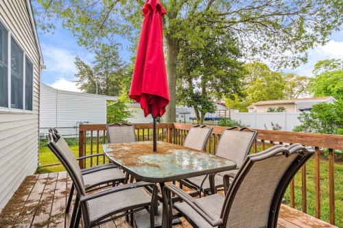 A wooden deck with a glass table and eight chairs, featuring a red umbrella, surrounded by greenery.