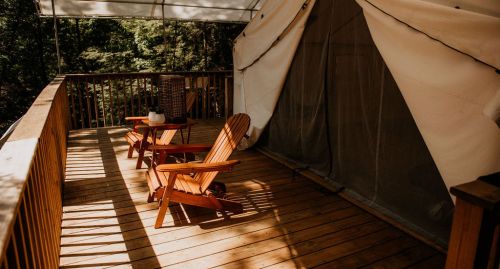 Two wooden Adirondack chairs on a deck in front of a tent, surrounded by trees and dappled sunlight.