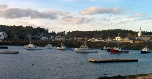 A scenic harbor with several boats anchored, surrounded by buildings and trees under a partly cloudy sky.