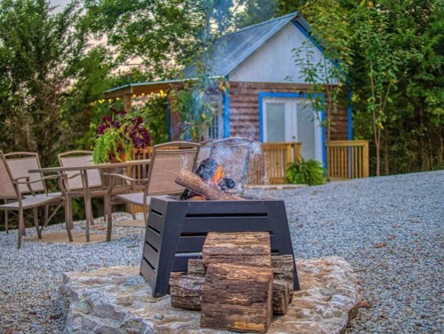 A cozy outdoor scene featuring a fire pit with logs, surrounded by gravel and a small cabin in the background.