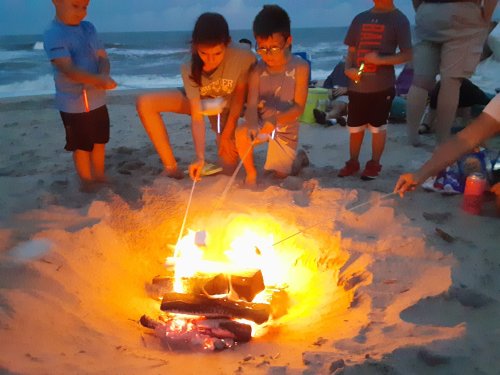 Children roasting marshmallows over a beach bonfire at dusk, with the ocean in the background.