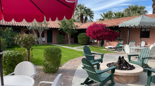 A sunny courtyard with green grass, a fire pit, and colorful plants, surrounded by adobe-style buildings and lounge chairs.