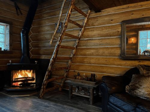Cozy cabin interior with a wooden ladder, fireplace, and leather couch, featuring rustic log walls and warm lighting.