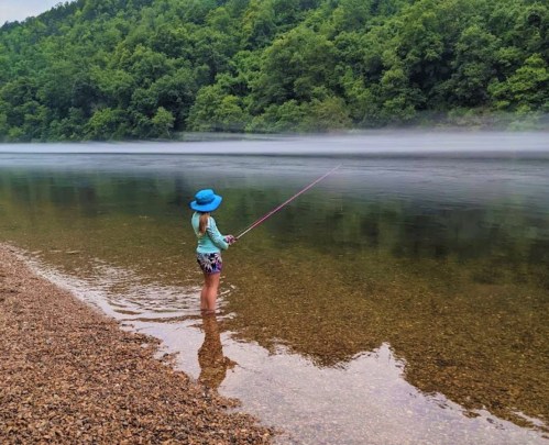 A child in a blue hat stands in shallow water, fishing by a misty river surrounded by lush green trees.