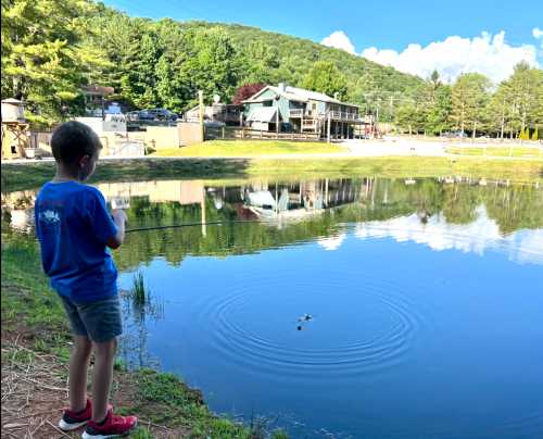 A child fishing by a calm pond, with trees and a building reflected in the water under a blue sky.