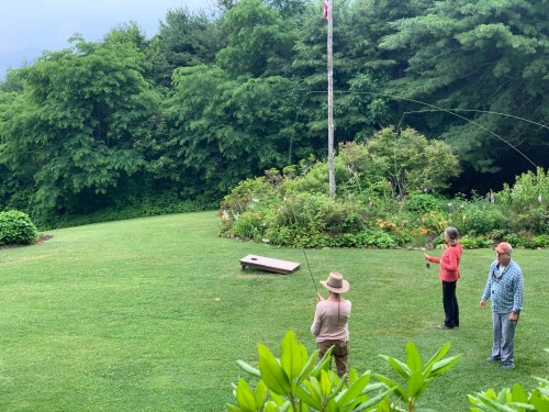 Three people fishing in a lush green garden with a flagpole and colorful flowers in the background.