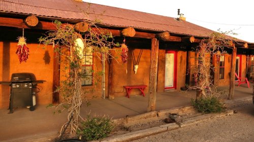 Adobe-style building with a red roof, hanging chili peppers, and a grill on a porch, surrounded by desert vegetation.