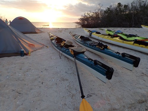 Kayaks on a sandy beach at sunset, with tents and trees in the background.