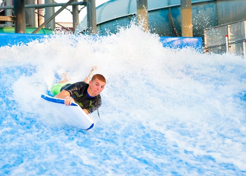 A person rides a bodyboard on a wave at a water park, splashing water around them.